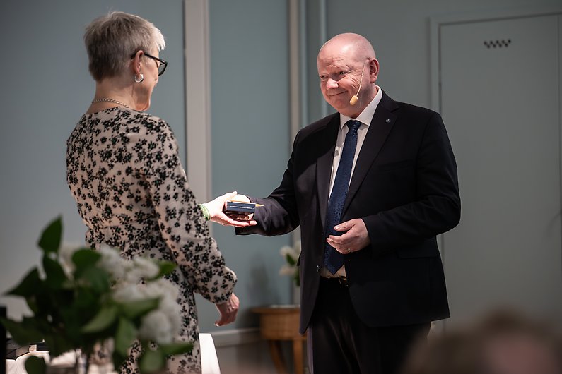 Rektor Hans Adolfsson räcker över gåva till kvinnlig person som ses bakifrån