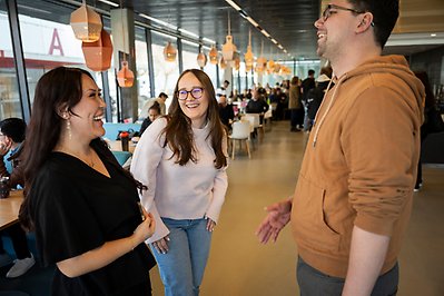 Tre personer samtalar glatt. I bakgrunden sitter människor vid bord och studerar.