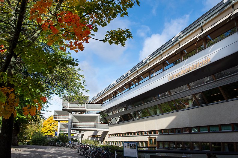 Foto på del av universitetsbiblioteket med skylten "Universitetsbibliotek" på fasaden. Till vänster i bild syns flera träd med klara höstfärger.